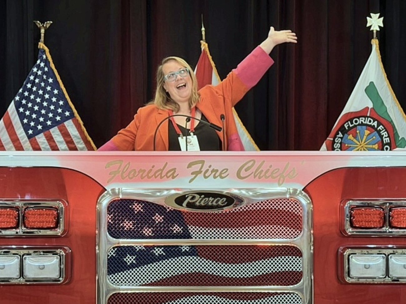 Laurie Sharp-Page on stage at the Florida Fire Chiefs' Association conference, arms raised behind a fire-truck podium